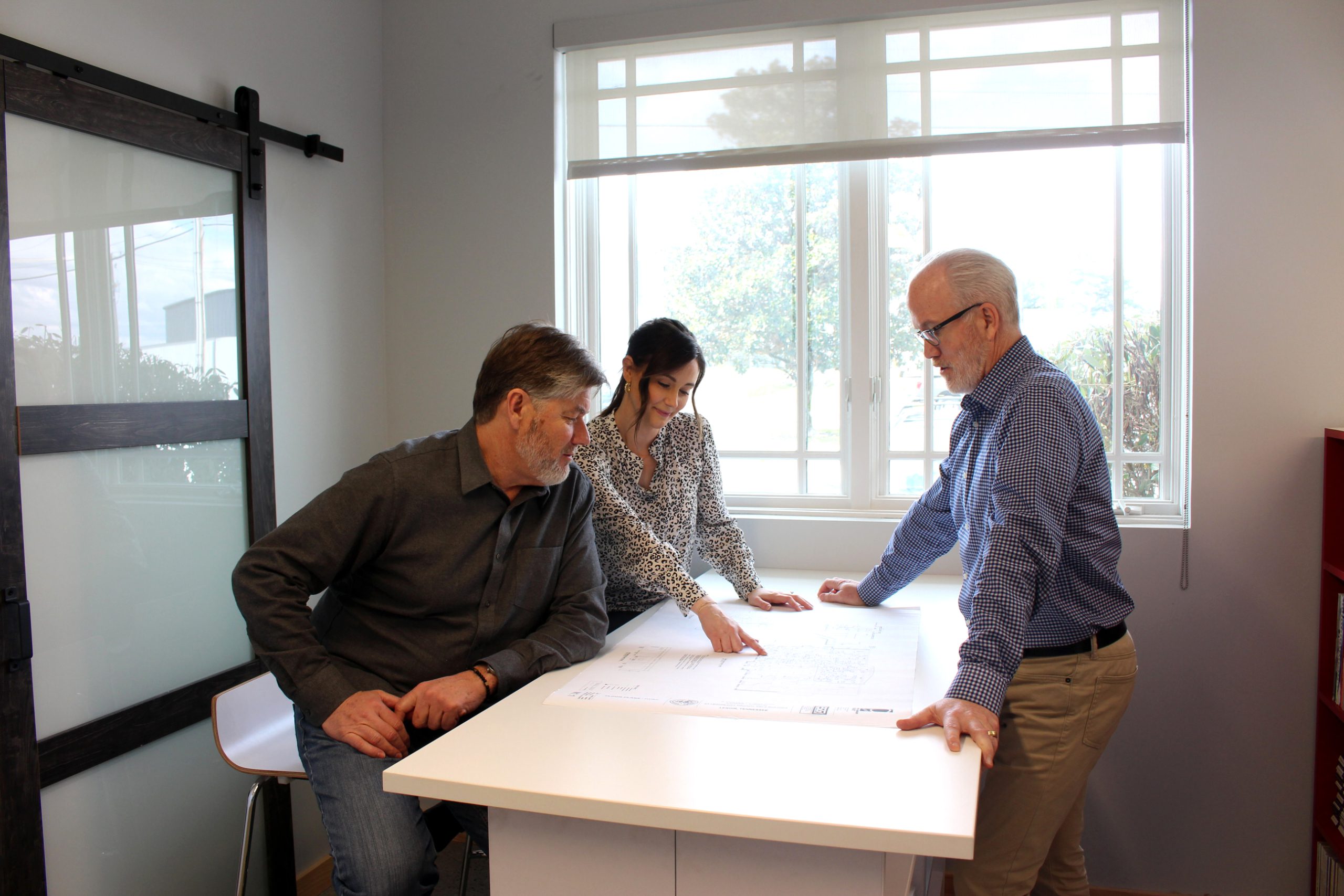 Three architects collaborating over architectural plans in a bright office setting, discussing design elements and career opportunities in commercial architecture.
