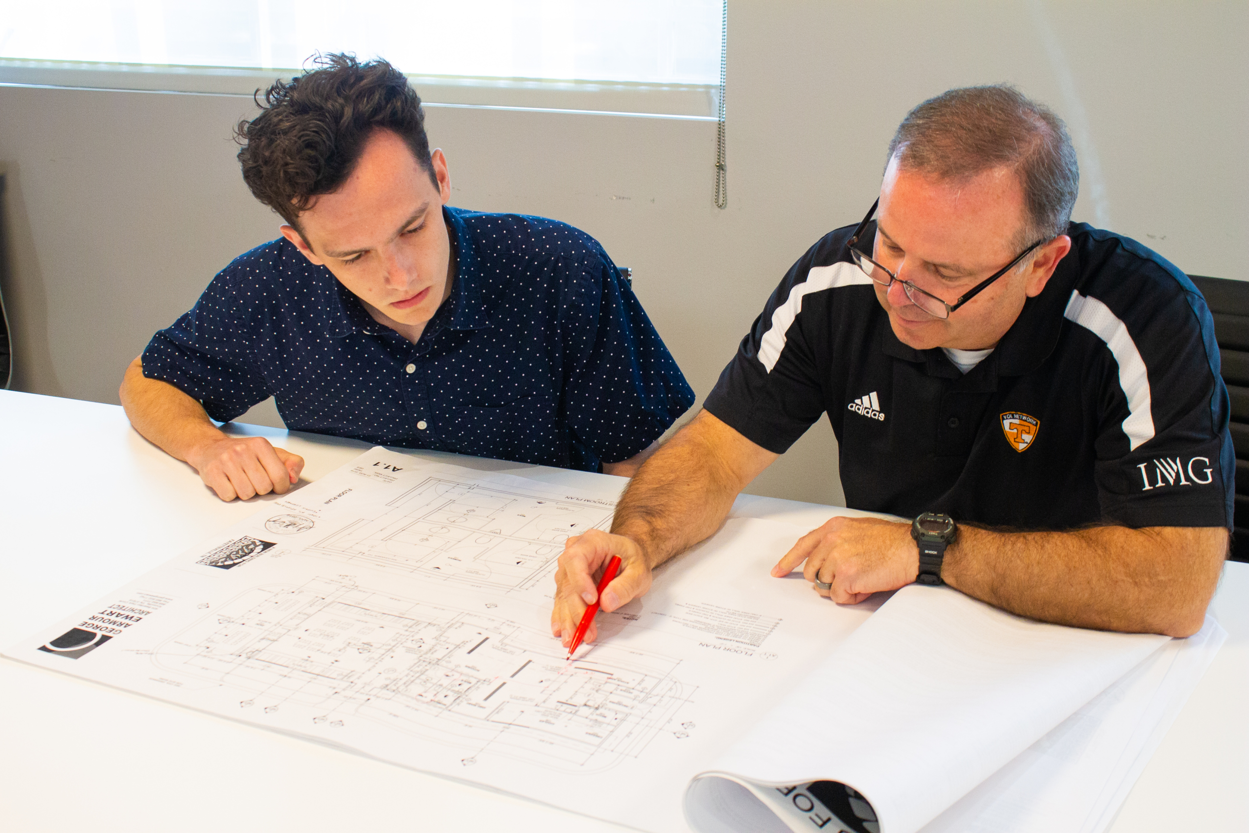 Two men reviewing architectural plans at a table, one pointing with a red pen, emphasizing collaboration in architecture and design careers.