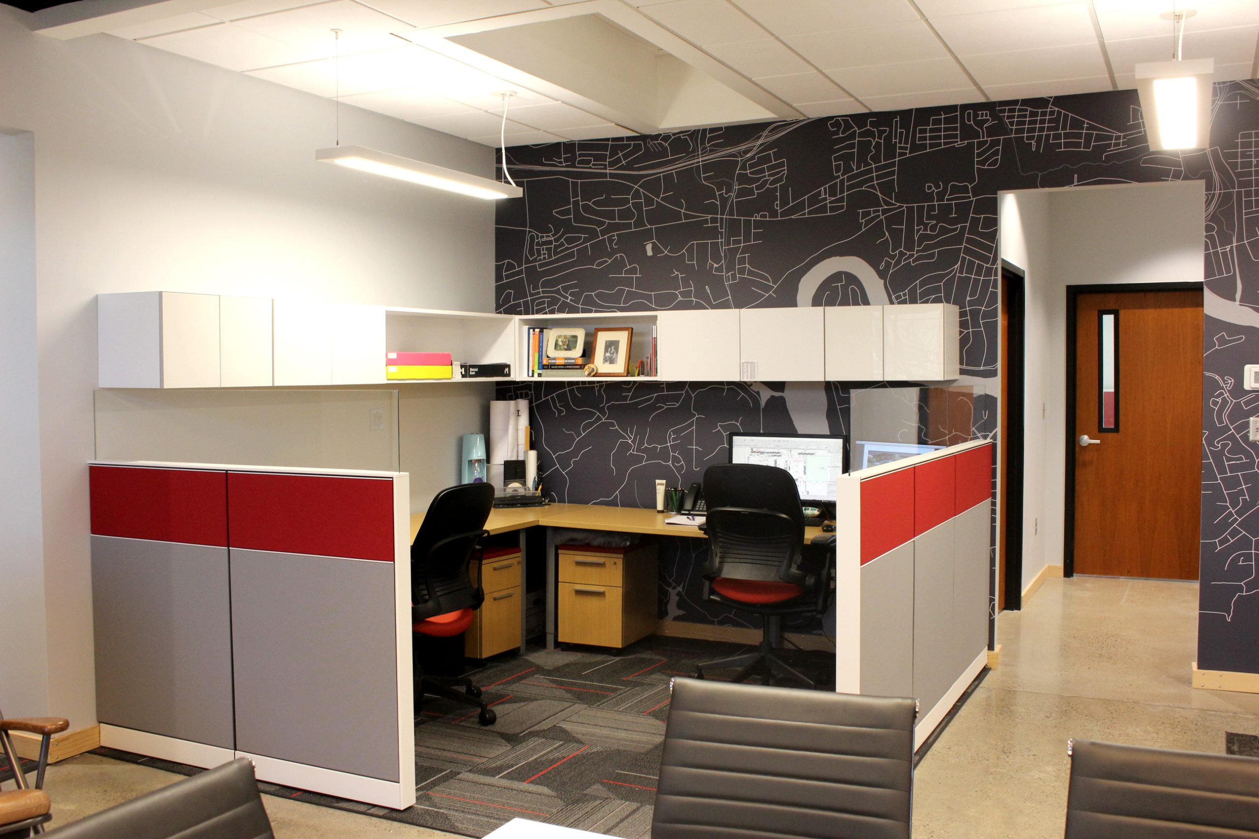 Modern architectural office workspace with dual desks, red and gray cabinetry, and a map-patterned wall, reflecting GAE Architecture + Design's collaborative project management environment.