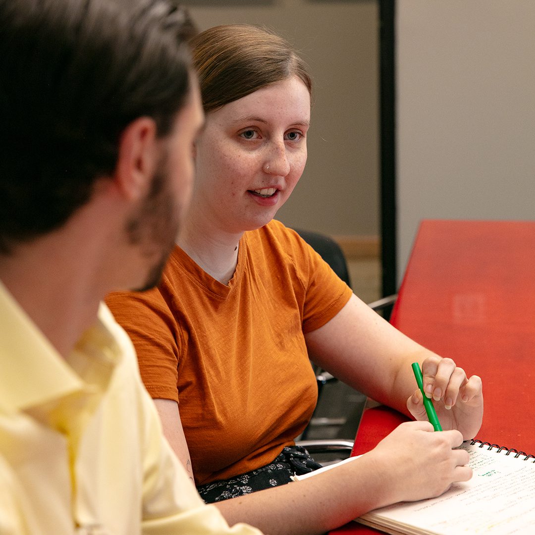 Aubrey White, Architectural Associate at GAEA, engaged in discussion with a colleague, holding a green pen and taking notes in a notebook on a red table, reflecting the firm's collaborative and creative environment.