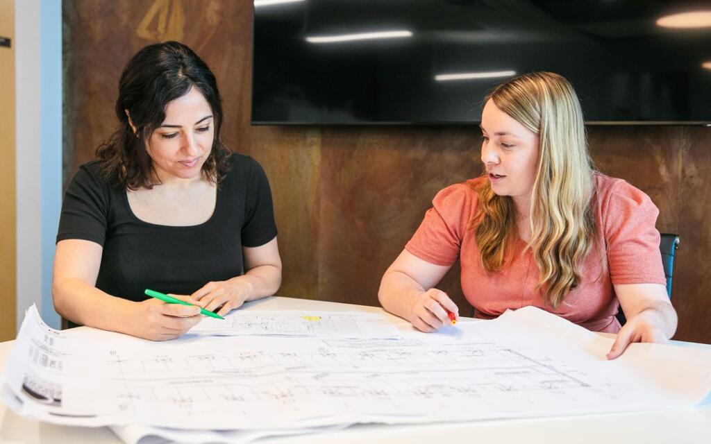 Two women collaborating on architectural blueprints, discussing design elements and project details, in a modern office setting, emphasizing teamwork in architecture and design.