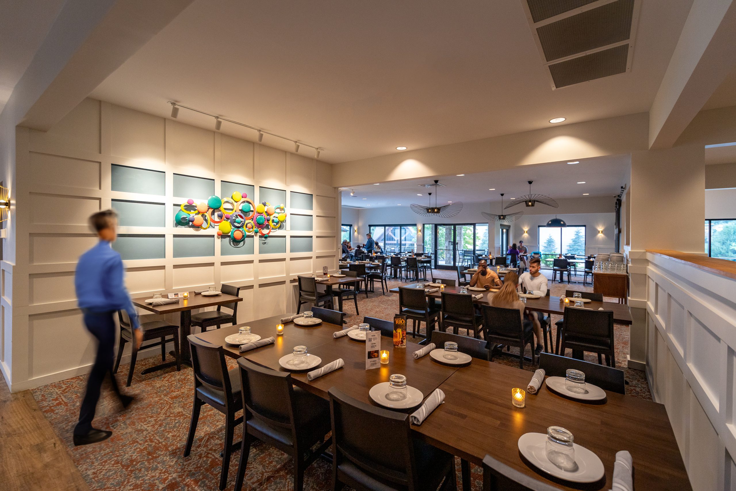 Interior dining space of Pizza Rio, featuring a colorful wall art installation, wooden tables set with plates and candles, and a waiter in a blue shirt, designed and built by George Ewart, a top commercial architect firm in Knoxville, TN.