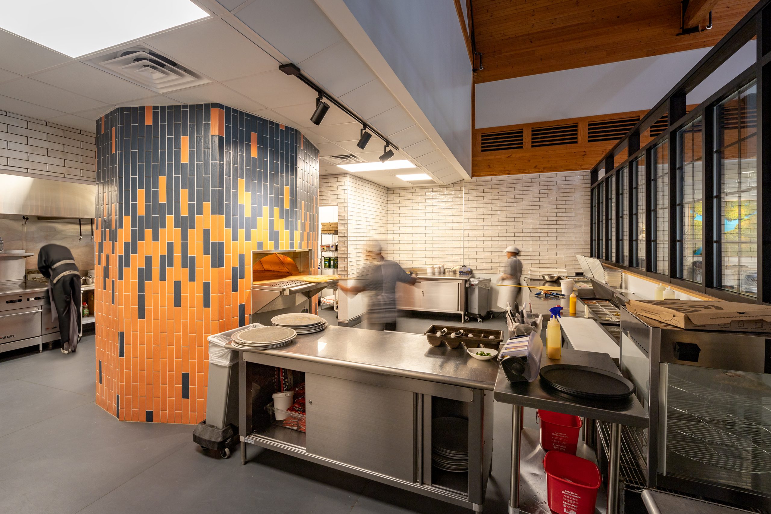 Kitchen interior of Pizza Rio featuring a vibrant tile pizza oven with orange and navy patterns, stainless steel appliances, and staff preparing food, designed and built by George Ewart, a top commercial architect firm in Knoxville, TN.