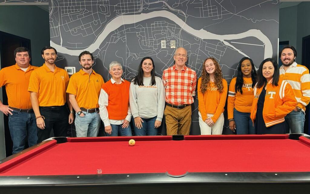 Group of GAE Architecture + Design team members in orange attire, posing in front of a map backdrop and a red pool table, representing a collaborative and vibrant work culture in Knoxville, TN.