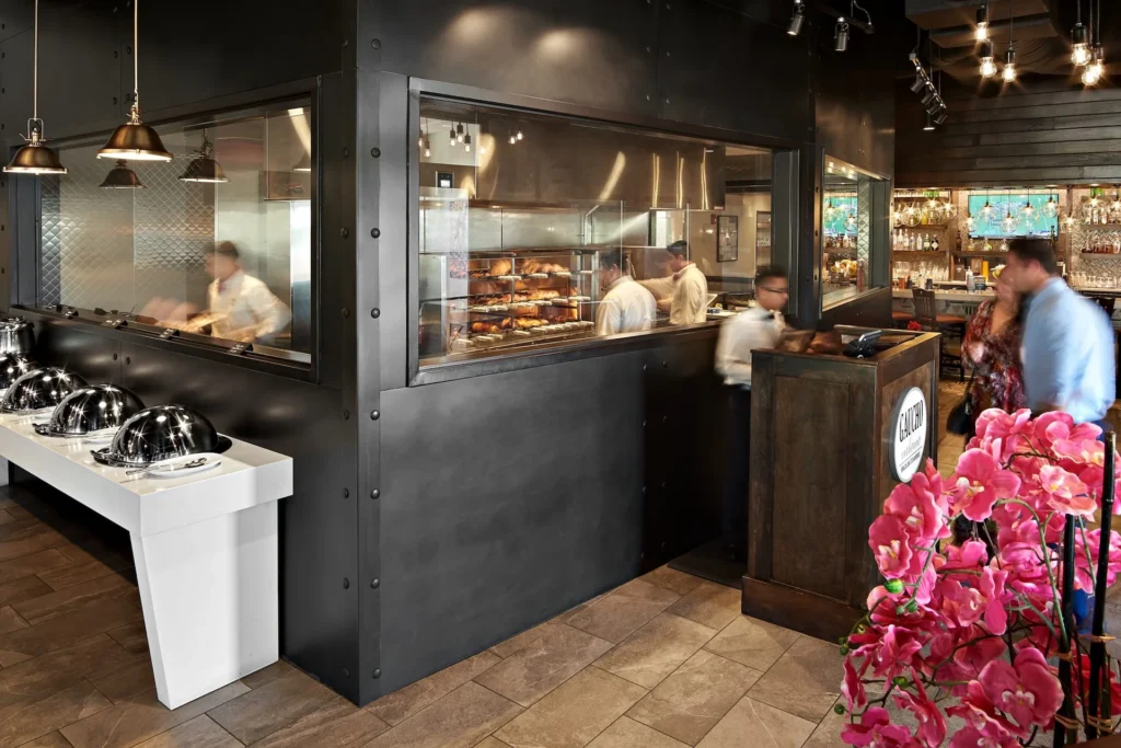 Interior view of Gaucho Urbano restaurant in Pigeon Forge, showcasing a modern dining space with a glass wall revealing the kitchen, chefs preparing meats, and a buffet setup, designed and built by George Ewart, a top commercial architect firm in Knoxville, TN.
