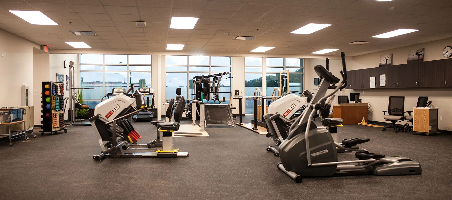 Physical therapy gym at CHRISTUS Health St. Mary’s Outpatient Center featuring exercise equipment, rehabilitation tools, and natural light from large windows, designed and built by George Ewart, a top commercial architect firm in Knoxville, TN.