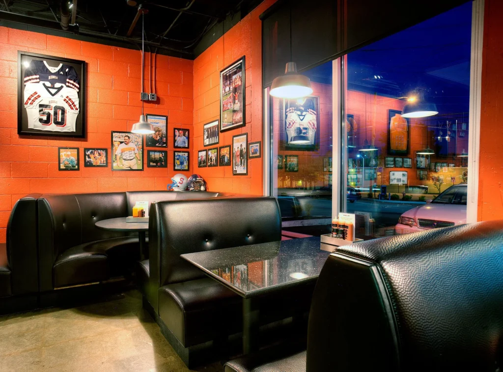 Cozy interior of Dead End BBQ restaurant featuring black leather booths, a round table, and vibrant orange walls adorned with sports memorabilia and framed photographs, designed and built by George Ewart, a top commercial architect firm in Knoxville, TN.