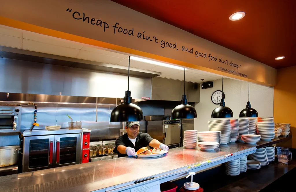 Chef serving barbecue dish in open kitchen at Dead End BBQ, with quote about food quality visible above, emphasizing transparency and community connection in Knoxville, TN, designed and built by George Ewart, a top commercial architect firm in Knoxville, TN.