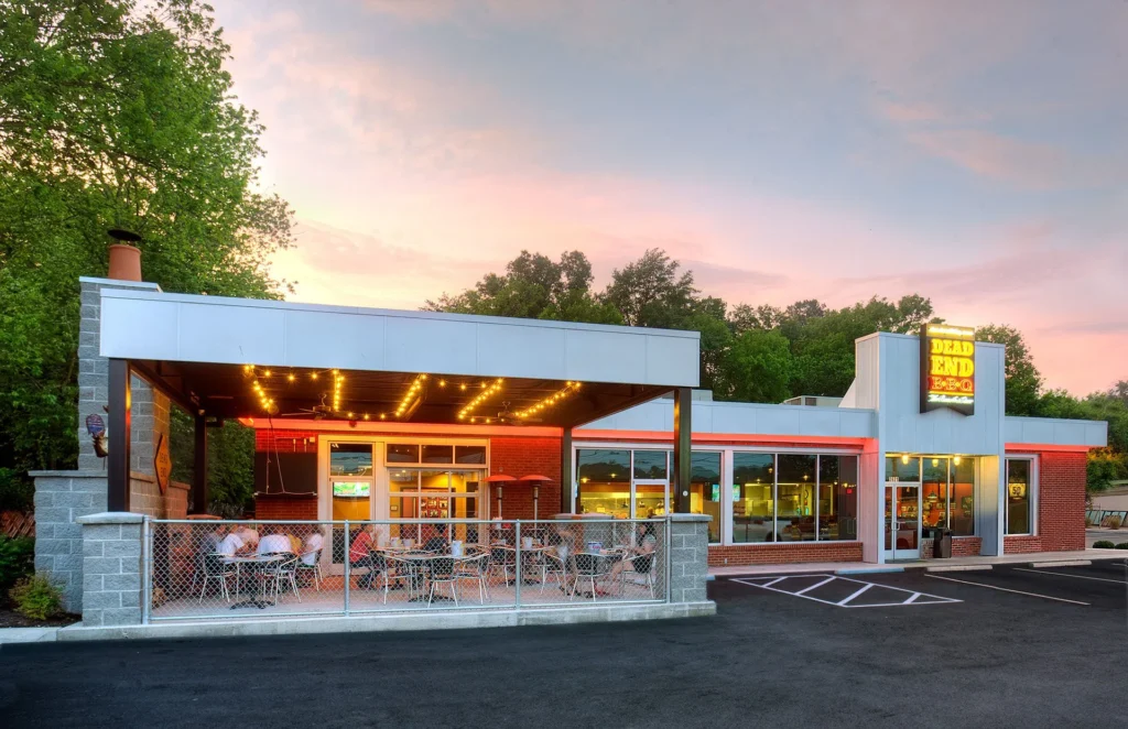 Exterior view of Dead End BBQ restaurant in Knoxville, TN, featuring a modern design with outdoor seating, illuminated patio, and large windows showcasing the dining area, designed and built by George Ewart, a top commercial architect firm in Knoxville, TN.