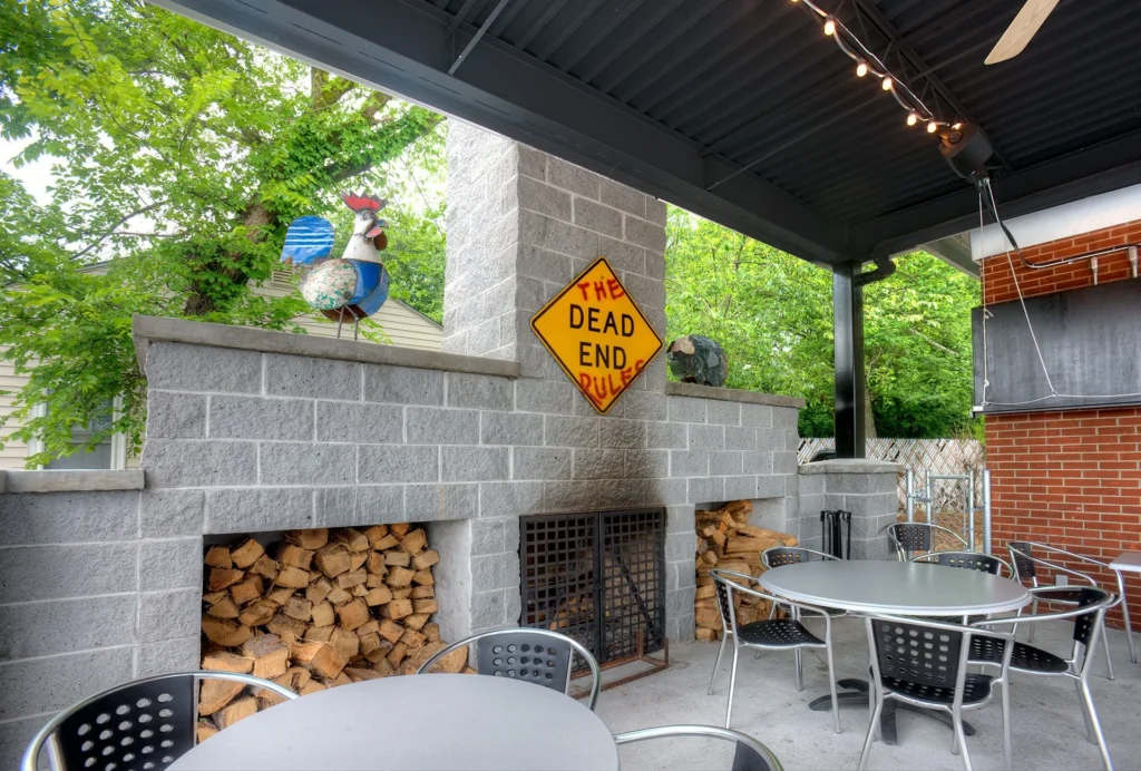 Outdoor dining area at Dead End BBQ, featuring a stone wall with stacked firewood, a whimsical rooster sculpture, and a sign reading "THE DEAD END RULES," designed to foster community connection, reflecting the restaurant's role as a neighborhood gathering place in Knoxville, TN, designed and built by George Ewart, a top commercial architect firm in Knoxville, TN.