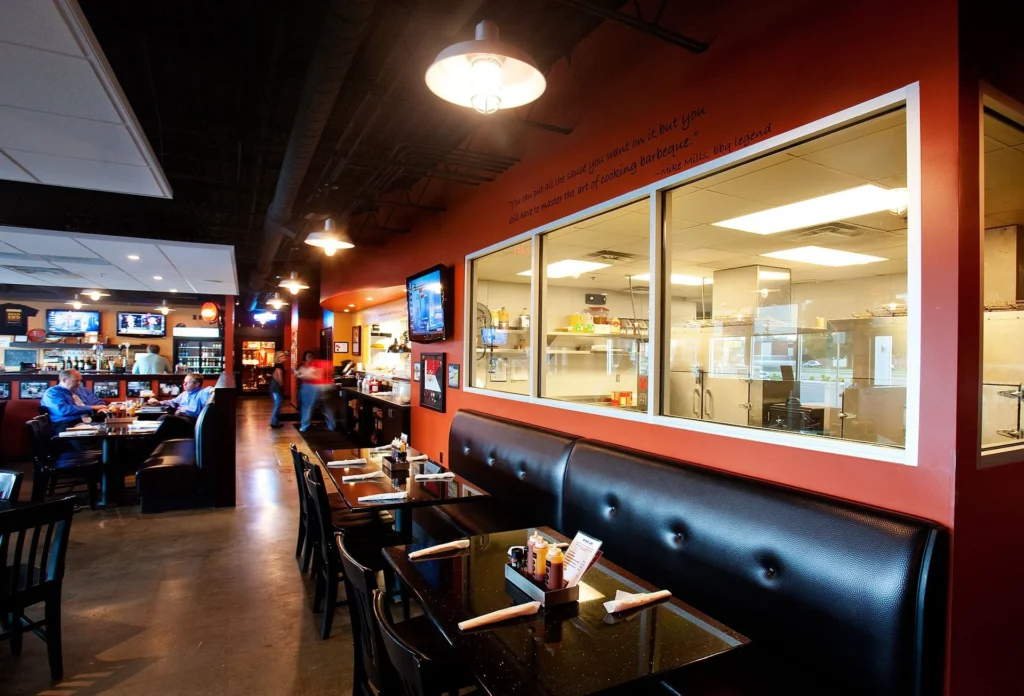 Interior view of Dead End BBQ restaurant in Knoxville, TN, featuring dining area with black booths, tables set for guests, and a visible smoke room showcasing pitmasters at work, emphasizing community gathering and transparency in design, created by commercial architects.