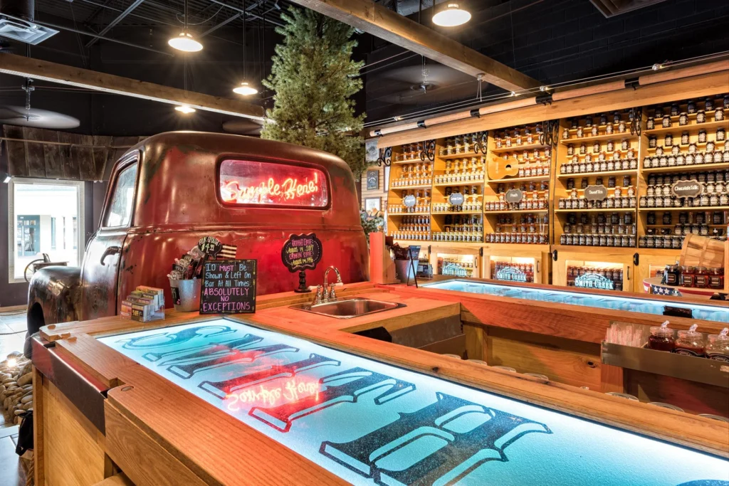 Vintage pickup truck front integrated as a communal tasting bar with illuminated glass countertops, surrounded by rustic wood finishes and shelves of moonshine jars, reflecting the Smoky Mountains' outdoor theme in the Smith Creek Moonshine retail space, designed and built by George Ewart, a top commercial architect firm in Knoxville, TN.