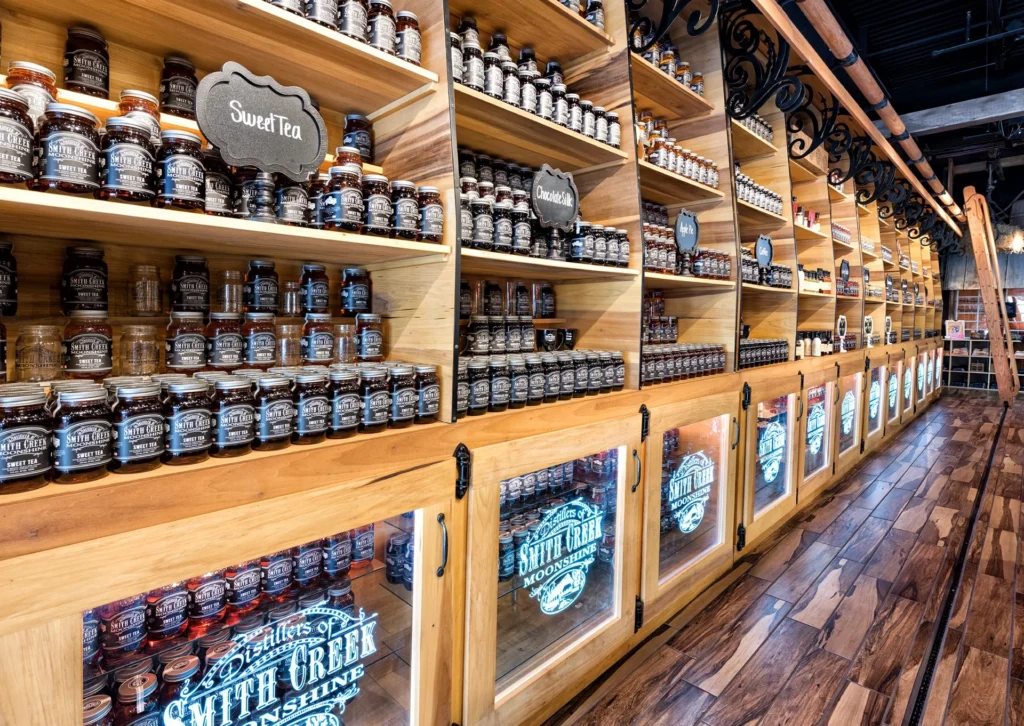 Interior display of Smith Creek Moonshine retail space featuring wooden shelves filled with various jars of moonshine, including "Sweet Tea" and "Chocolate Silk," with illuminated glass cabinets and rustic wood finishes, designed and built by George Ewart, a top commercial architect firm in Knoxville, TN.