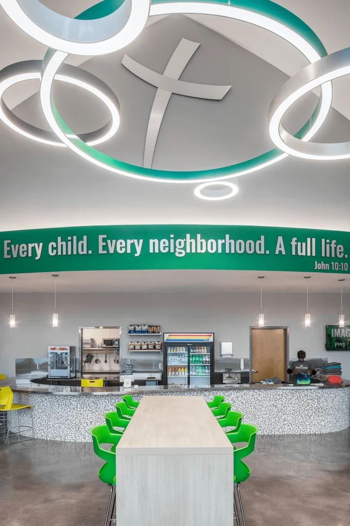 Interior view of a community center café featuring a long table with bright green chairs, modern lighting fixtures, and a wall displaying the phrase "Every child. Every neighborhood. A full life." designed and built by George Ewart, a top commercial architect firm in Knoxville, TN.