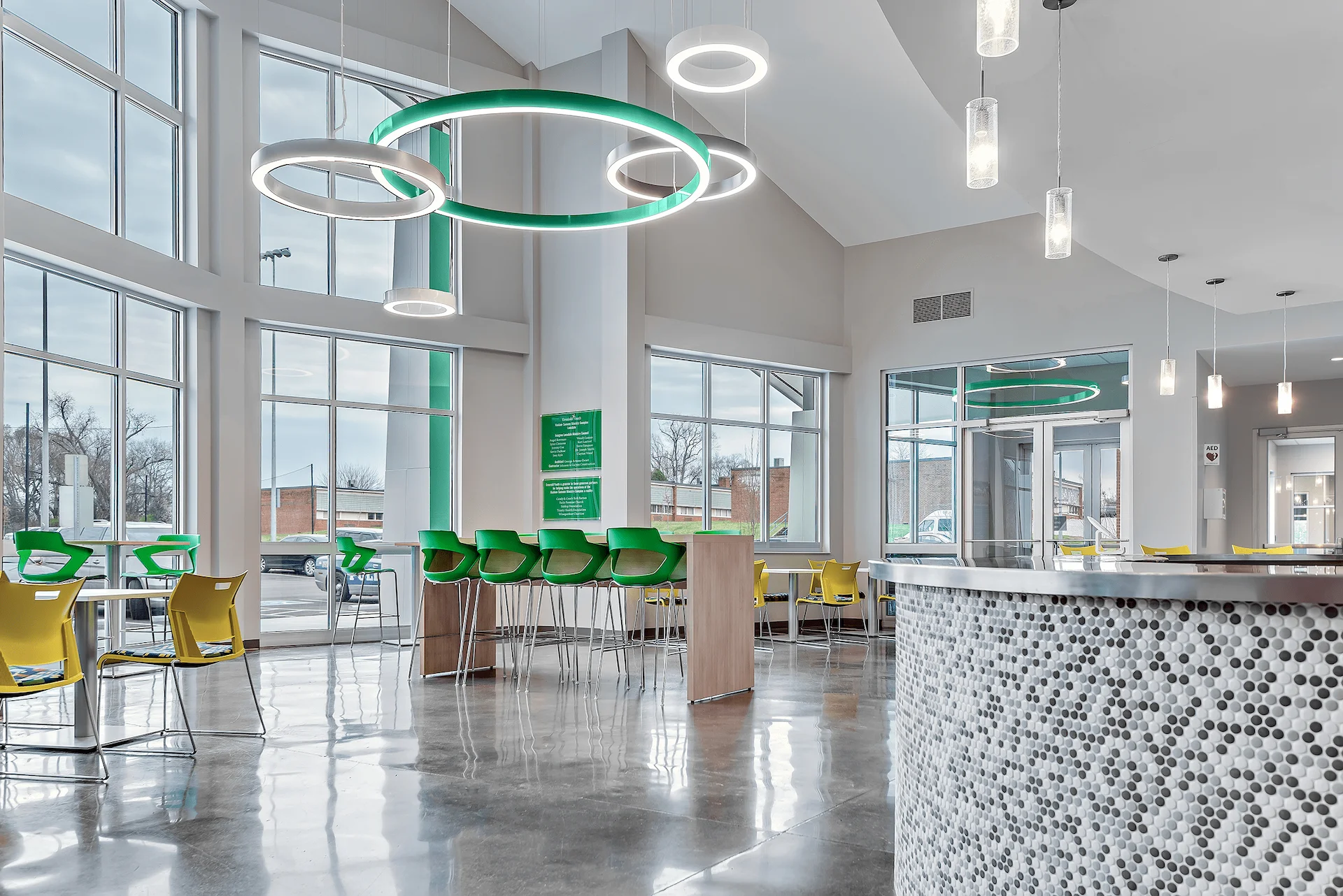 Bright, modern interior of the Emerald Haslam Sansom Ministry Complex in Knoxville, featuring circular light fixtures, green and yellow seating, large windows, and a polished concrete floor, designed for community engagement and youth development.