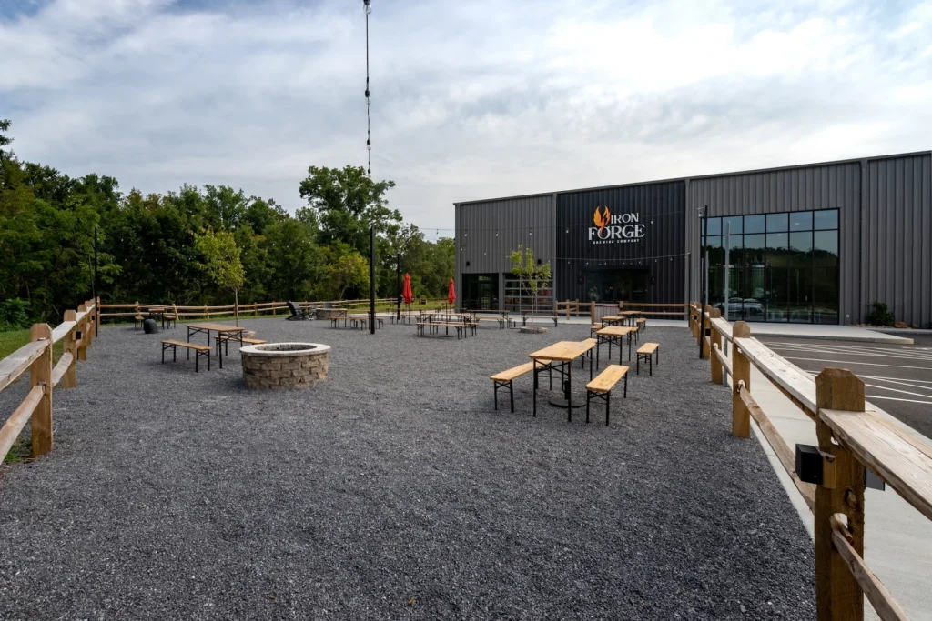 Outdoor seating area at Iron Forge Brewing Co. featuring gravel surface, wooden tables, and fire pit, with brewery building in the background, designed and built by George Ewart, a top commercial architect firm in Knoxville, TN.
