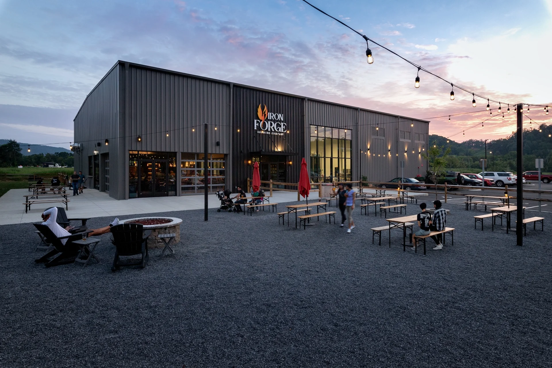 Iron Forge Brewing Co. exterior featuring a modern design by GAE Architecture + Design, outdoor seating area with picnic tables and fire pit, illuminated by string lights at dusk, located in Knoxville, TN.