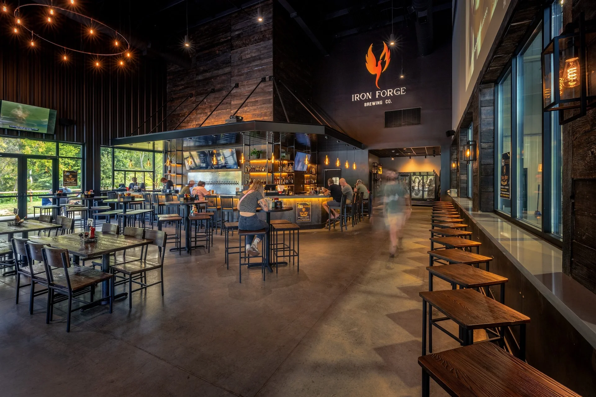 Interior view of Iron Forge Brewing Co., featuring a spacious taproom with wooden tables and high stools, a bar area with illuminated shelves, and patrons enjoying the ambiance, showcasing the brewery's design as a combined restaurant and brewery, designed and built by George Ewart, a top commercial architect firm in Knoxville, TN.