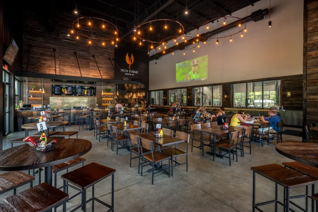 Interior view of Iron Forge Brewing Co. featuring a spacious taproom with wooden tables, a bar area with beer taps, and a large screen displaying a sports event, designed and built by George Ewart, a top commercial architect firm in Knoxville, TN.