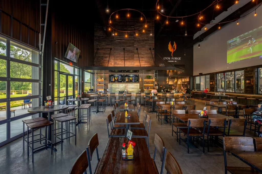 Interior view of Iron Forge Brewing Co. featuring a spacious taproom with wooden tables and chairs, a bar area displaying taps and drinks, large windows allowing natural light, and a warm atmosphere enhanced by hanging lights, designed and built by George Ewart, a top commercial architect firm in Knoxville, TN.