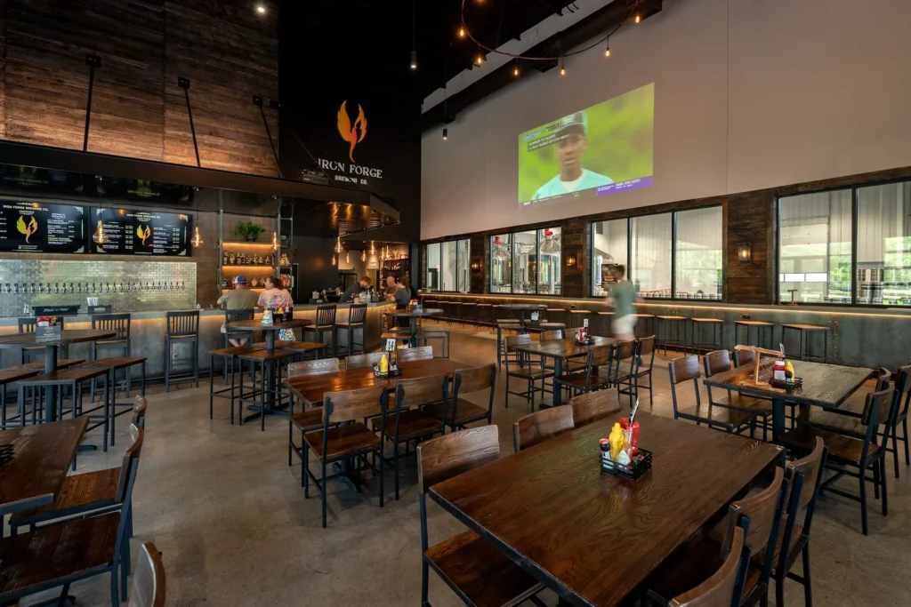 Interior view of Iron Forge Brewing Co. featuring a spacious taproom with wooden tables and chairs, a bar with multiple beer taps, and a large screen displaying a sports event, designed and built by George Ewart, a top commercial architect firm in Knoxville, TN.