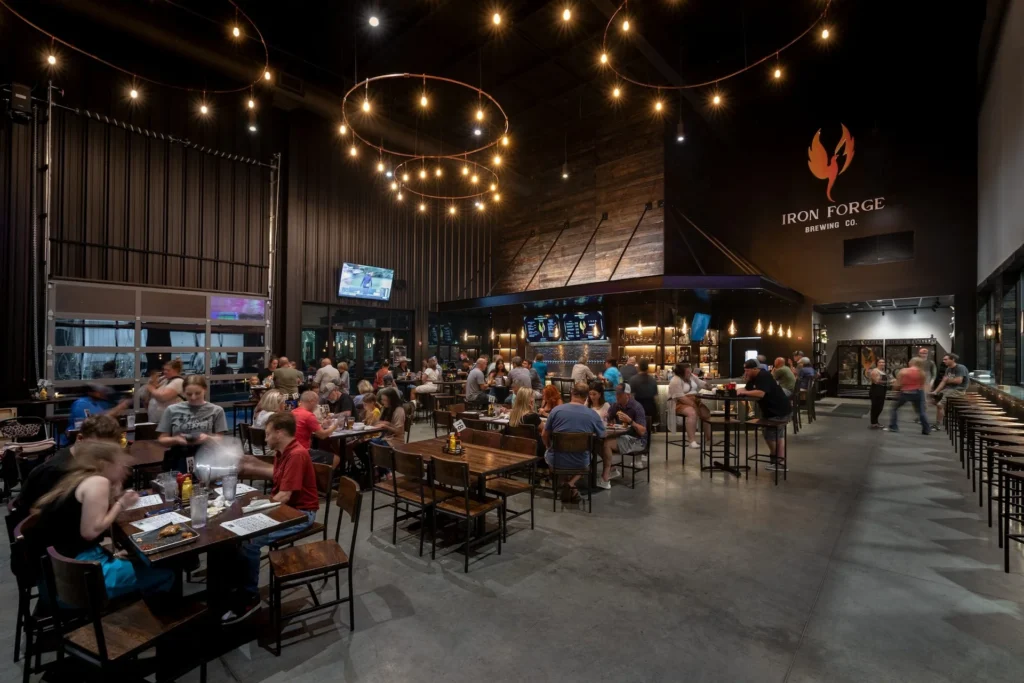 Interior view of Iron Forge Brewing Co. featuring a bustling taproom with patrons dining and socializing, illuminated by decorative lighting; designed and built by George Ewart, a top commercial architect firm in Knoxville, TN.