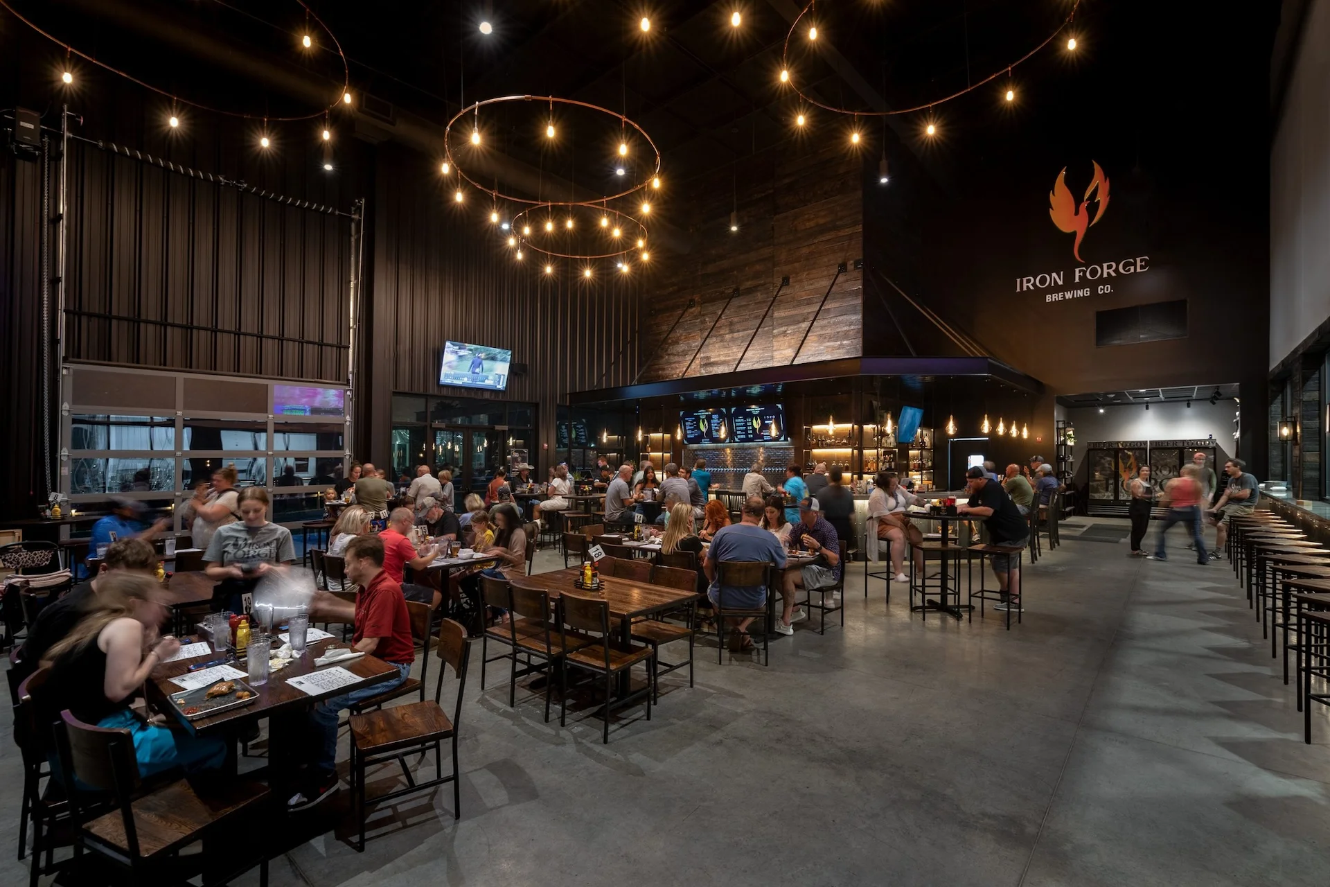 Interior of Iron Forge Brewing Co. featuring a bustling taproom with patrons dining and socializing, illuminated by pendant lights and showcasing a bar area with beer taps, designed and built by George Ewart, a top commercial architect firm in Knoxville, TN.