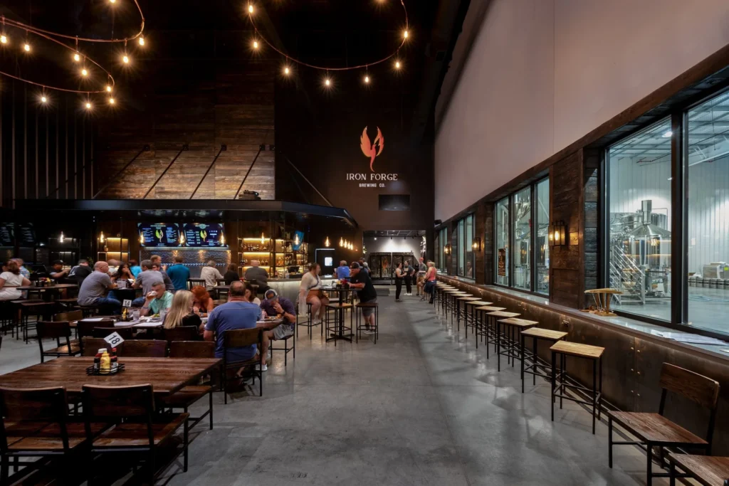 Interior of Iron Forge Brewing Co. featuring a spacious taproom with patrons dining, a bar area with illuminated menus, and large windows showcasing beer fermenters, designed and built by George Ewart, a top commercial architect firm in Knoxville, TN.