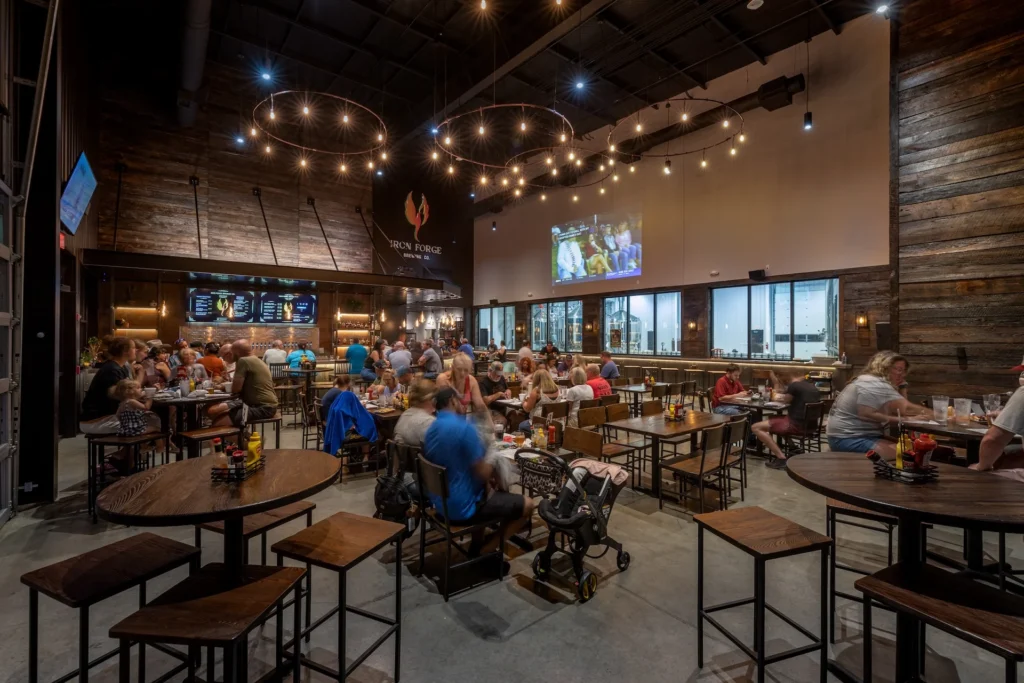 Interior view of Iron Forge Brewing Co. featuring a bustling taproom with patrons enjoying food and drinks, exposed brewing equipment visible behind glass, rustic wooden decor, and warm ambient lighting, designed and built by George Ewart, a top commercial architect firm in Knoxville, TN.
