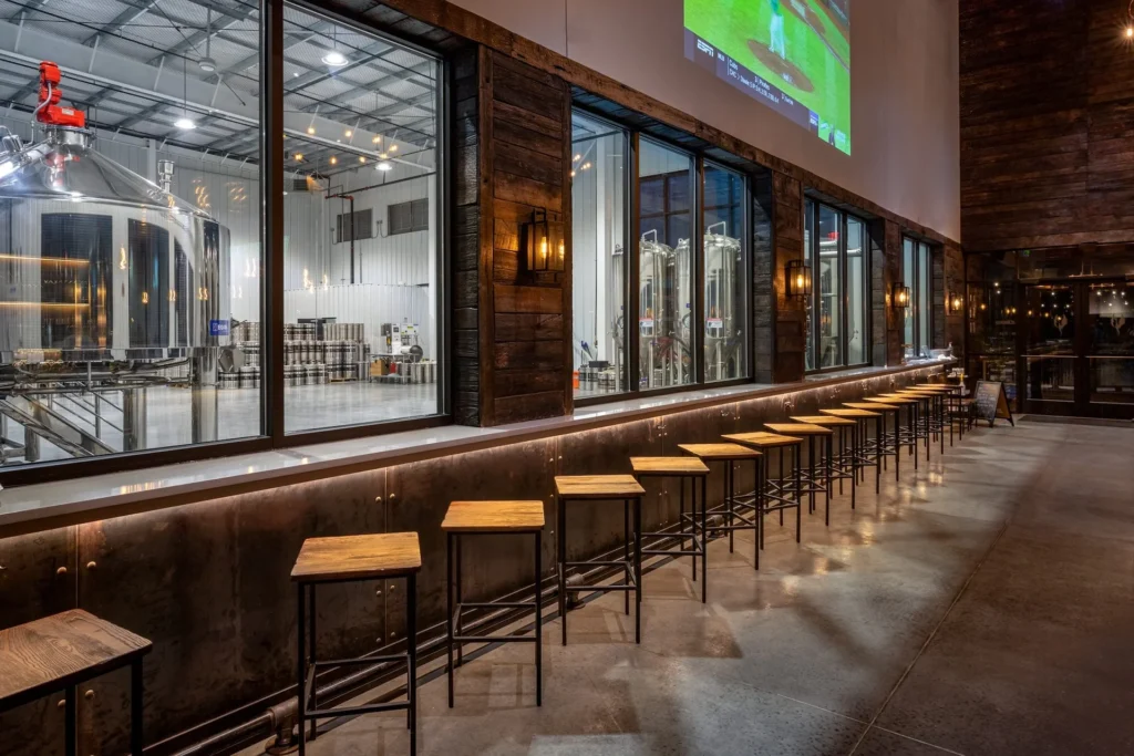 Interior view of Iron Forge Brewing Co. featuring a modern taproom with wooden bar stools, large windows showcasing beer fermenters, and a projector screen displaying sports, designed and built by George Ewart, a top commercial architect firm in Knoxville, TN.