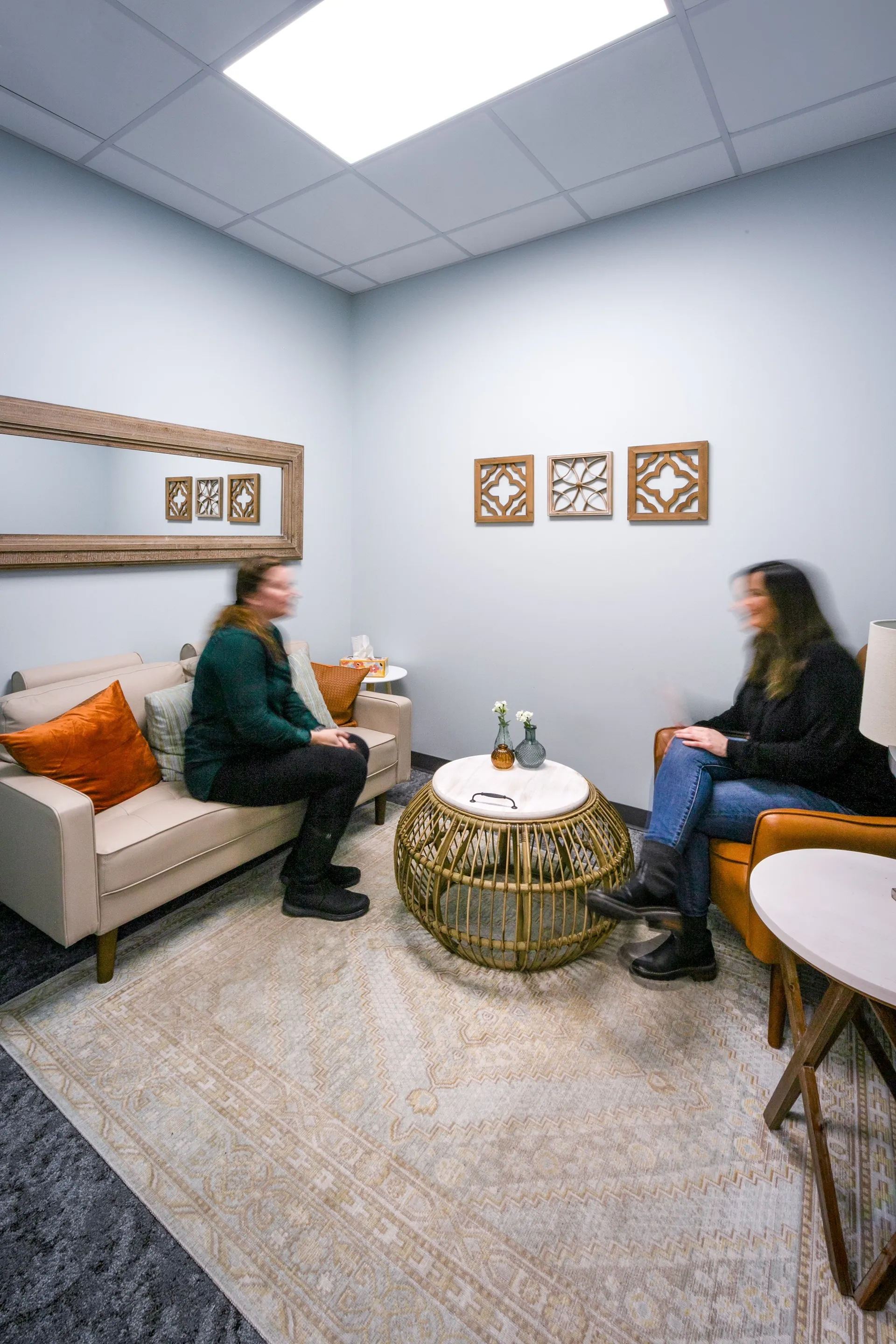 Interior consultation space at Catholic Charities featuring two women seated on a sofa and chair, decorative wooden wall art, and a central coffee table, emphasizing a welcoming environment for service delivery.