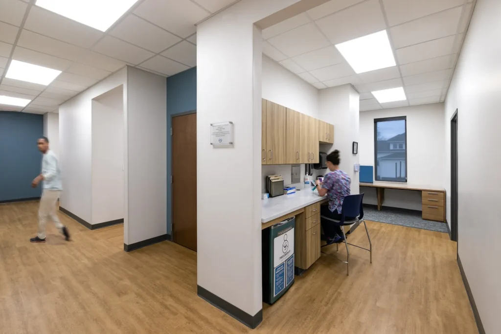 Interior view of renovated facility for Catholic Charities in Knoxville, TN, featuring a shared workspace with a woman at a desk and a man walking in the hallway, designed and built by George Ewart, a top commercial architect firm in Knoxville, TN, highlighting improved workflow and service delivery.