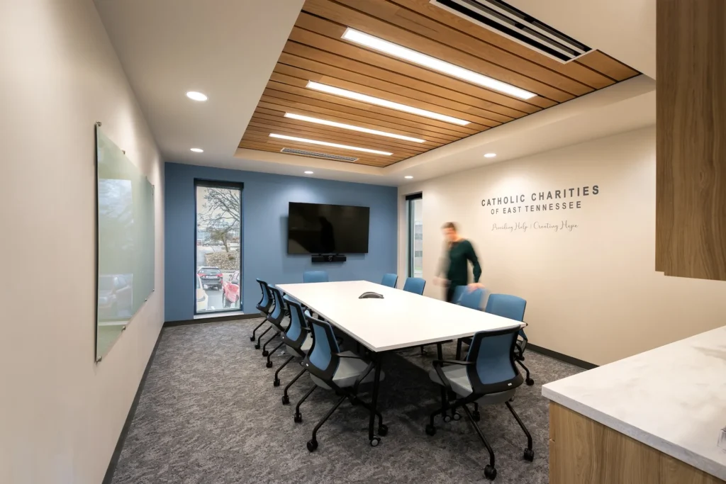 Modern conference room at Catholic Charities of East Tennessee featuring a large white table, blue chairs, wooden ceiling accents, and branding on the wall, designed and built by George Ewart, a top commercial architect firm in Knoxville, TN.