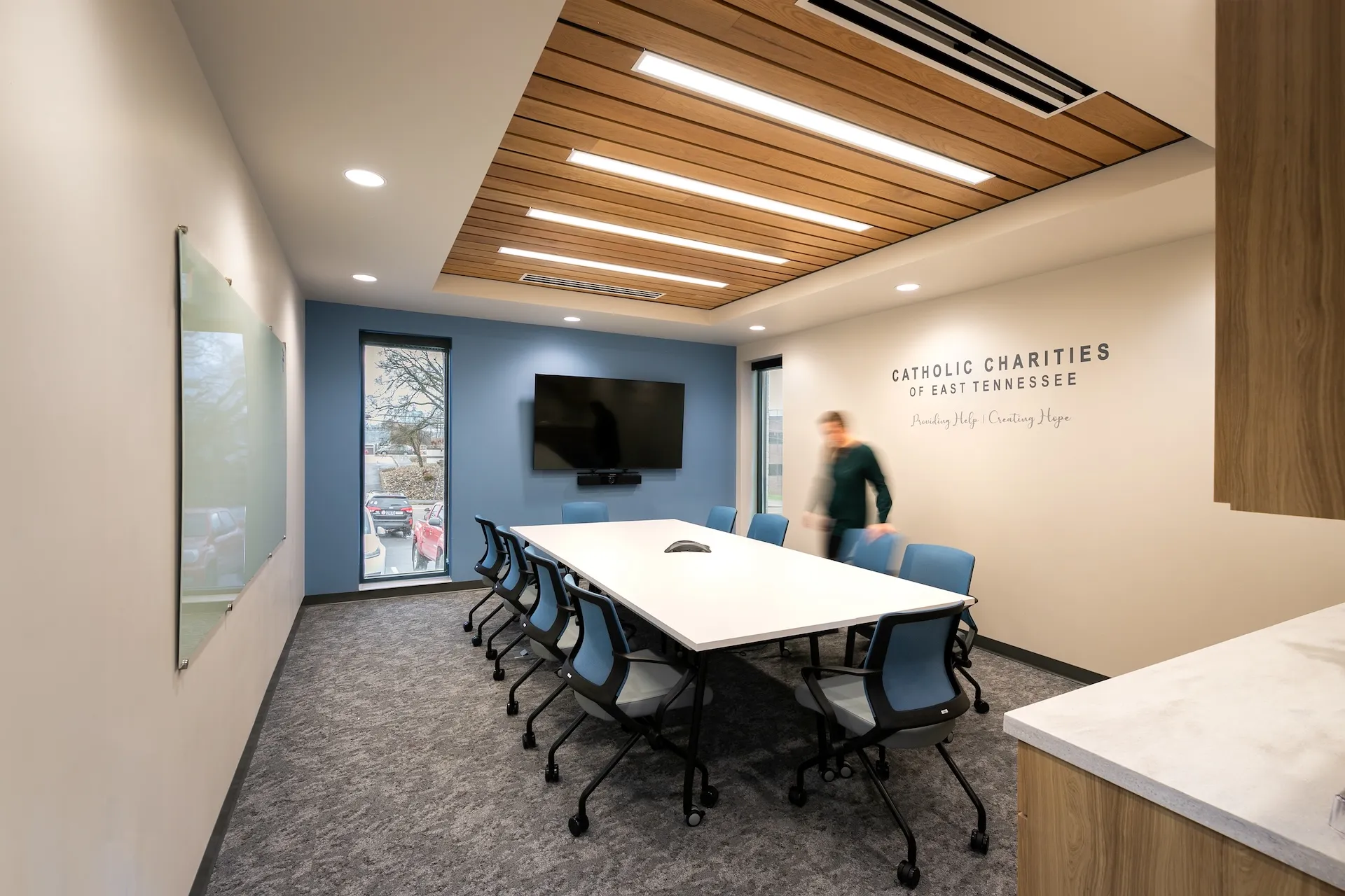 Conference room at Catholic Charities of East Tennessee featuring a large table, blue chairs, a wall-mounted TV, and branding text emphasizing community support.