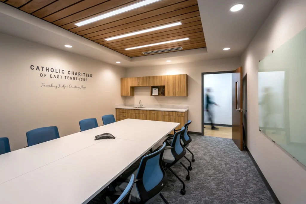 Conference room at Catholic Charities of East Tennessee featuring a long white table, blue chairs, wooden cabinetry, and a wall with the organization's name and mission statement, designed and built by George Ewart, a top commercial architect firm in Knoxville, TN.