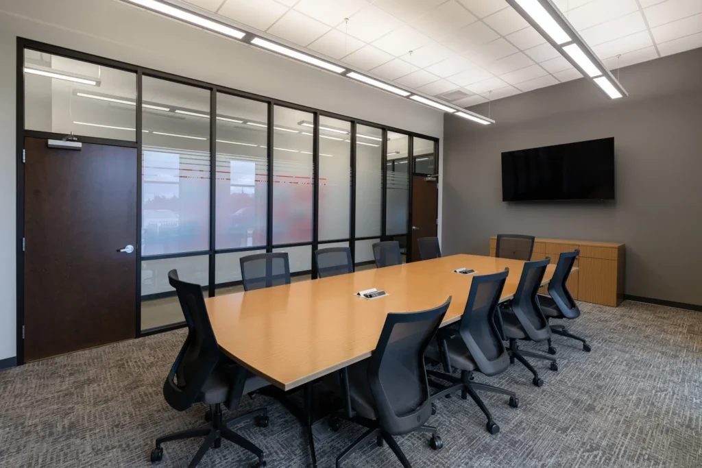 Modern conference room with a long wooden table, ergonomic black chairs, glass wall partitions, and a wall-mounted TV, designed for collaborative meetings in the Fraley & Schilling headquarters, a project by GAE Architecture + Design, commercial architects in Knoxville, TN.