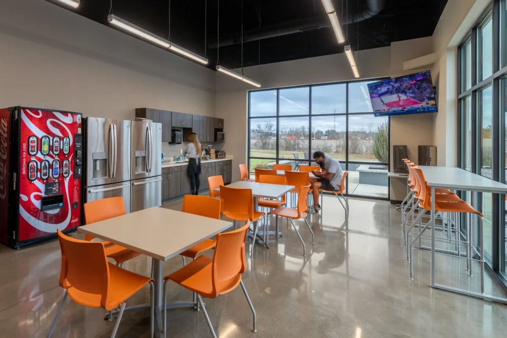 Modern office break room with orange chairs, vending machine, stainless steel appliances, large windows, and a TV, showcasing the collaborative environment designed for Fraley & Schilling's corporate headquarters in Knoxville, TN, by GAE Architecture + Design.