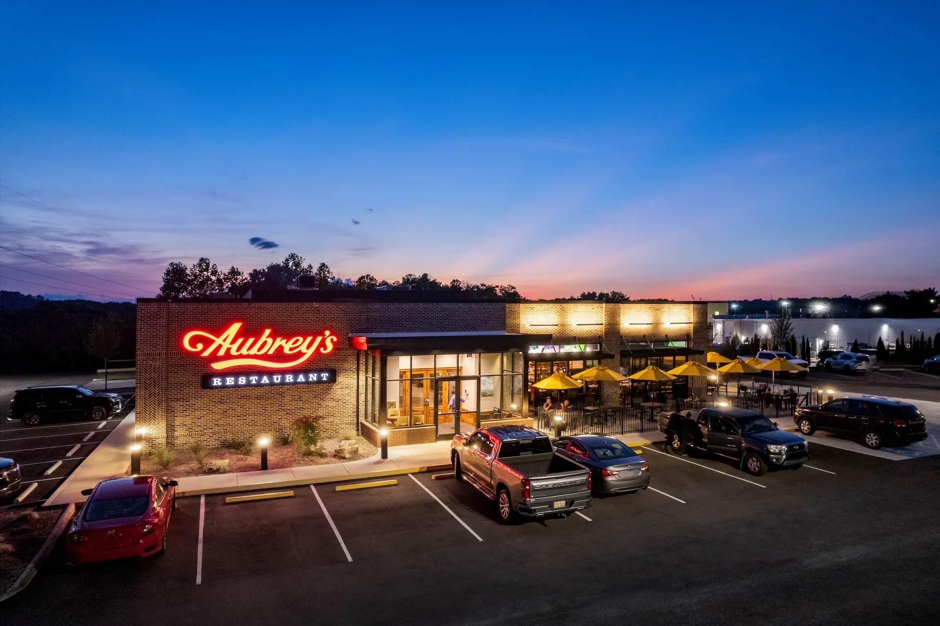 Aubrey's restaurant exterior at dusk, featuring illuminated signage, outdoor seating with yellow umbrellas, and a modern architectural design, showcasing the work of GAE Architecture + Design, commercial architects based in Knoxville, TN.