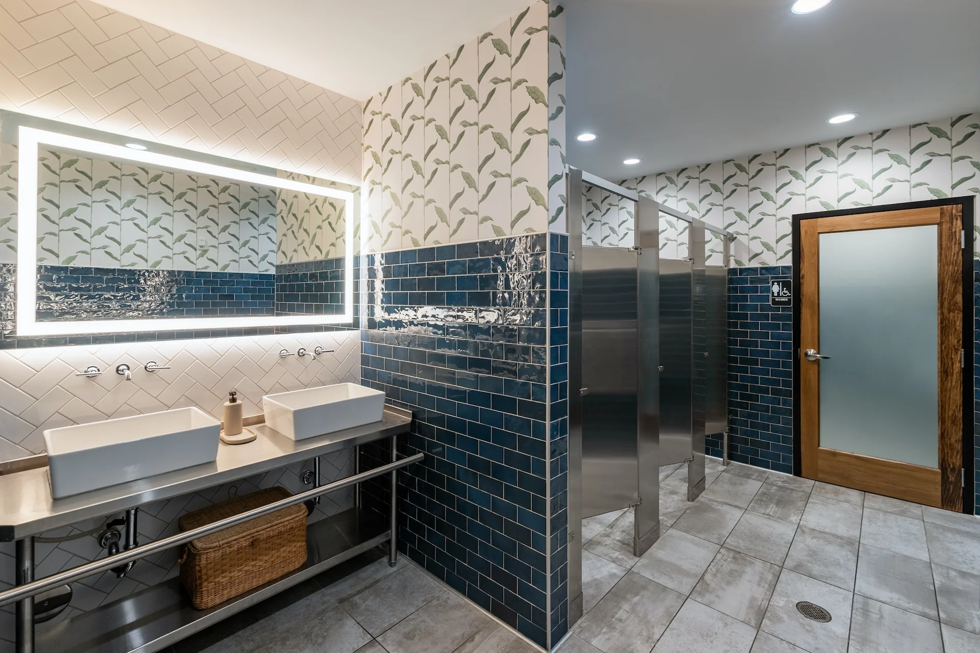 Modern restroom interior featuring dual vessel sinks, illuminated mirror, deep blue tiles, and botanical wallpaper, designed by GAE Architecture + Design for Aubrey's in Lenoir City, TN.