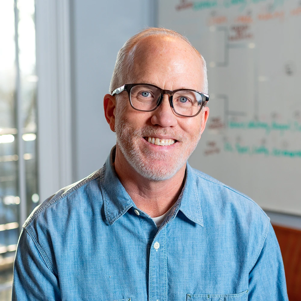 Stuart H. Anderson, project architect at GAE Architecture + Design, smiling in a casual blue shirt, with a whiteboard in the background featuring project notes, showcasing his approachable personality and professional environment in Knoxville, TN.